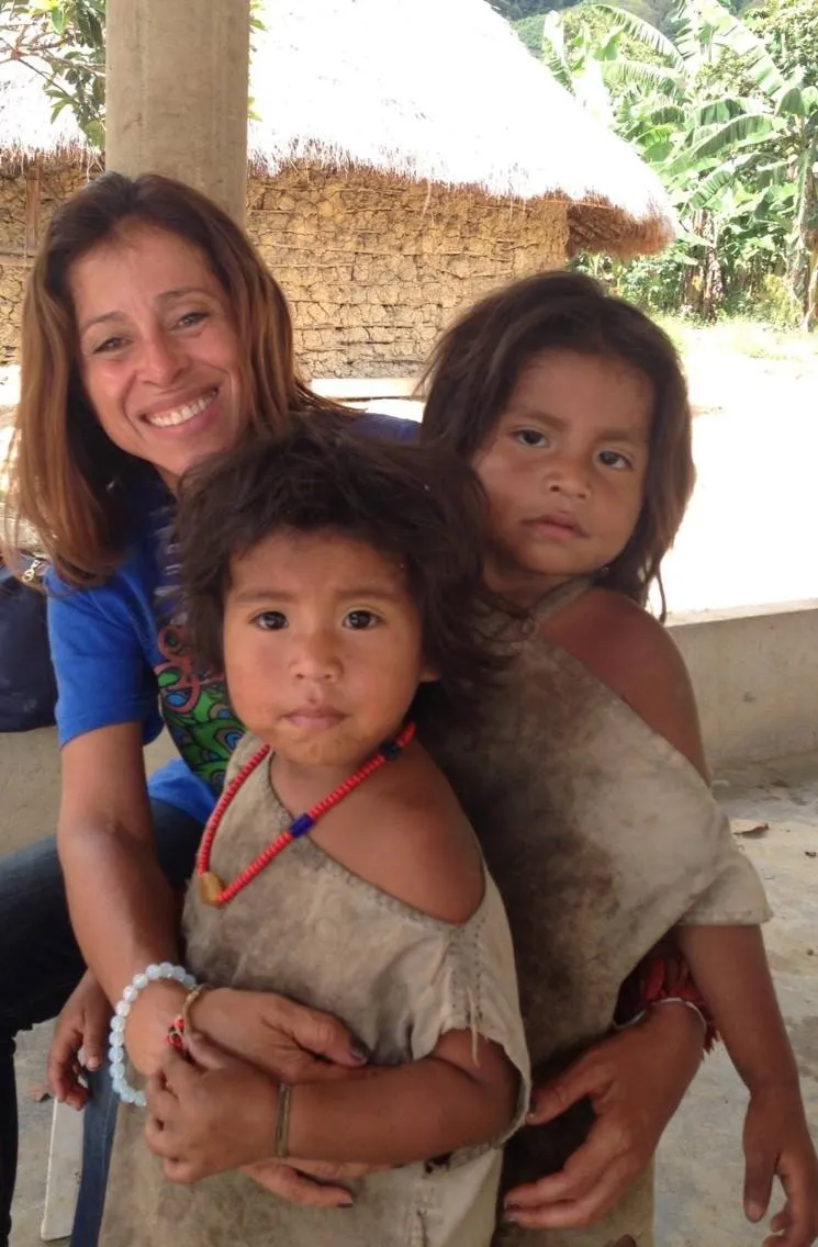 Claudia Aguirre, souriante, tient contre elle deux jeunes enfants kogis devant une case au toit de chaume, en Colombie.