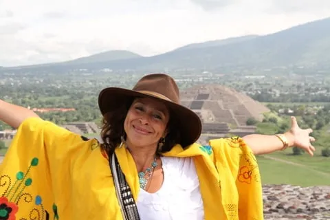 Claudia Aguirre, chapeau de feutre et châle jaune brodé, bras grands ouverts devant la pyramide de la Lune de Teotihuacán.