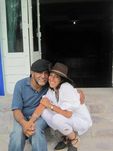 Claudia Aguirre, en blanc et chapeau, assise contre un homme sur le perron d'une maison ; tous deux rient.