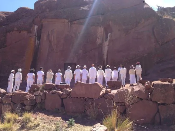 Le groupe, de dos et en blanc, face à la paroi rouge d'Aramu Muru ; un rai de lumière traverse l'image en diagonale.